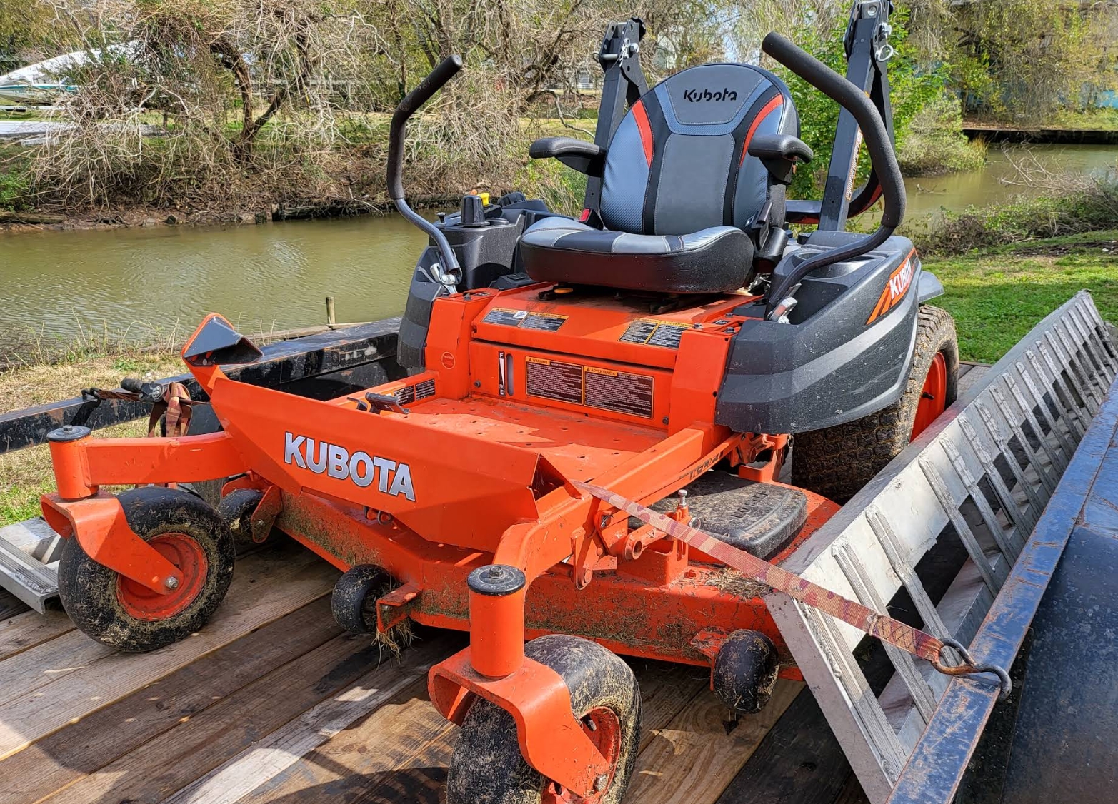 An orange zero-turn riding mower strapped down on a flatbed trailer beside a bayou, loading ramp still out