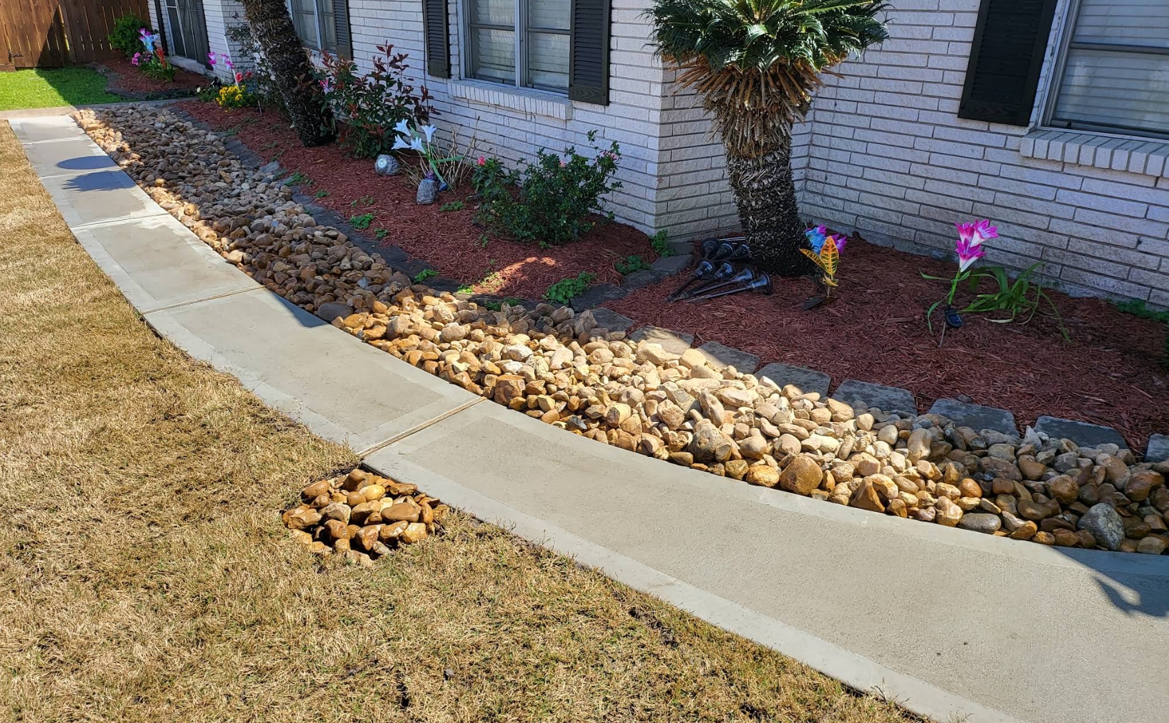 A river-rock bed over red mulch running along the front of a white brick home, with a sago palm and flowering plants